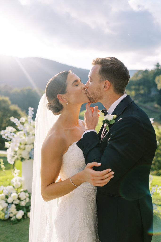 Bride and groom share a romantic kiss at their Lake Toxaway mountain-view ceremony near Asheville, surrounded by white florals and golden sunlight, planned by Two Sweet Sparrows.