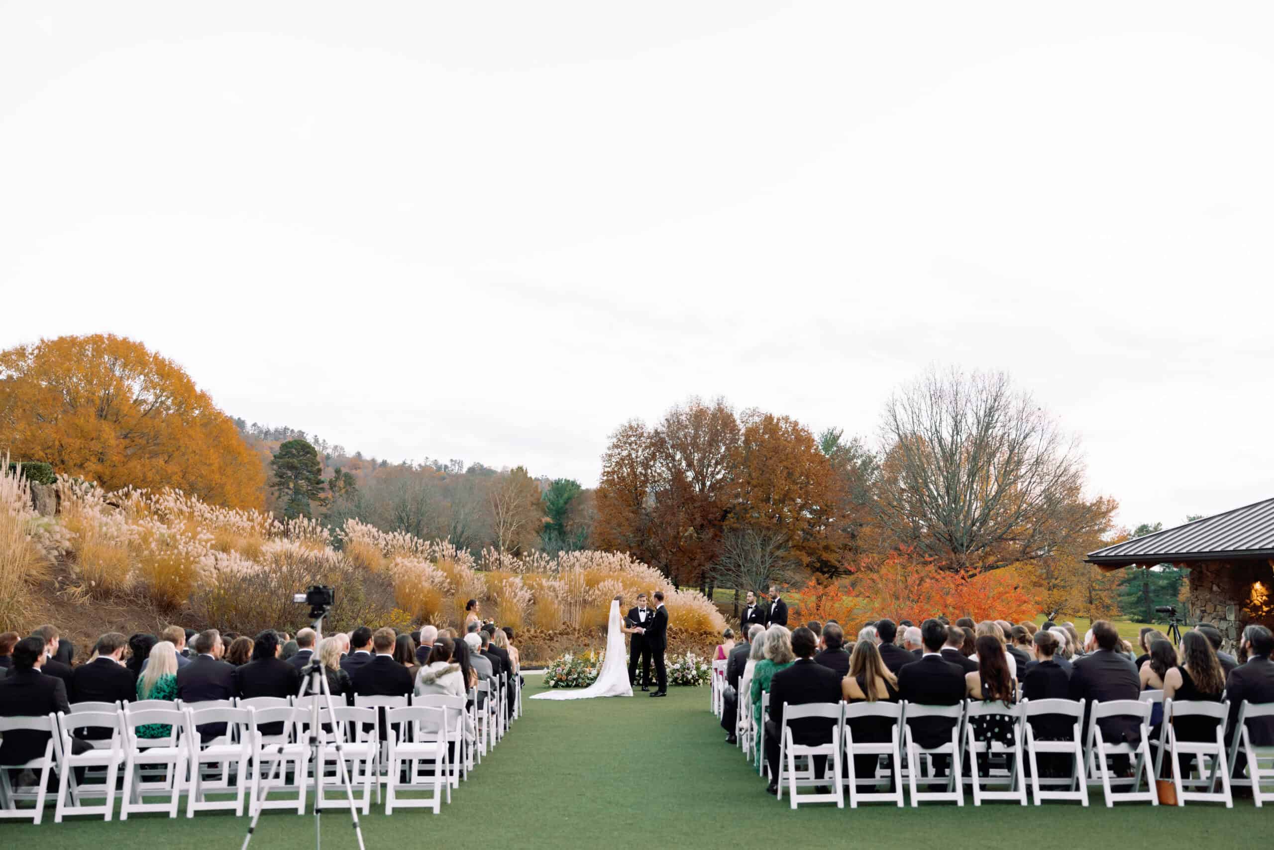 Outdoor wedding ceremony with guests seated on white chairs in fall setting.