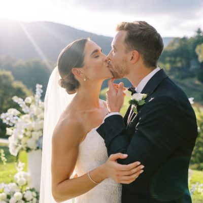 Bride and groom share a romantic kiss at their Lake Toxaway mountain-view ceremony near Asheville, surrounded by white florals and golden sunlight, planned by Two Sweet Sparrows.
